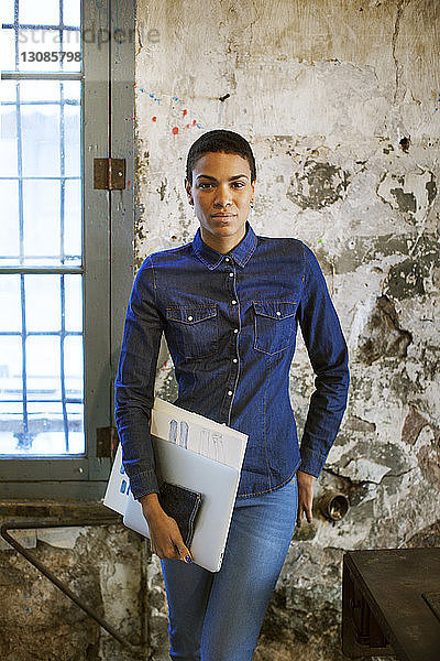Selbstbewusste Modedesignerin hält Dokumente in der Hand  während sie in einer alten Werkstatt steht