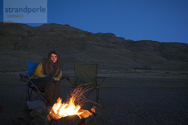 Porträt einer auf einem Stuhl sitzenden Frau am Lagerfeuer vor Bergen und blauem Himmel in der Alvord-Wüste
