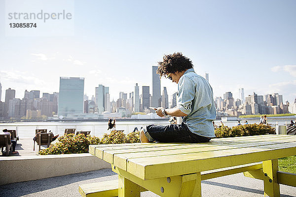 Junger Mann benutzt Tabletten  während er auf einer Bank gegen das Stadtbild sitzt