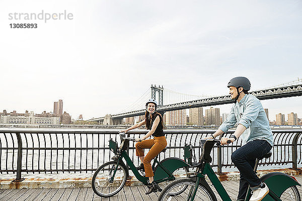 Glückliches Paar radelt auf Fußweg an der Manhattan-Brücke gegen den Himmel