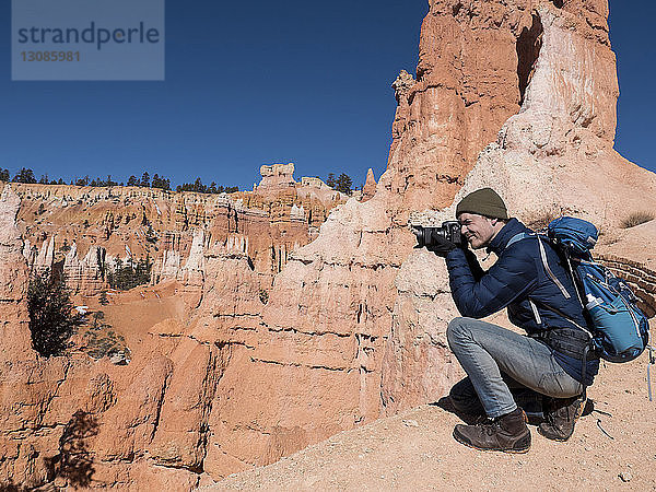 Seitenansicht eines Wanderers  der mit der Kamera fotografiert  während er in der Wüste vor Felsformationen kauert