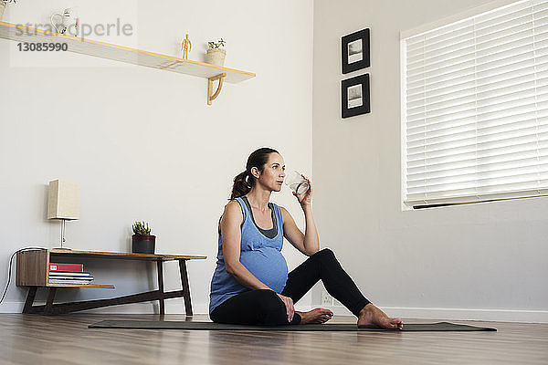 Schwangere Frau trinkt Wasser  während sie zu Hause auf der Gymnastikmatte sitzt
