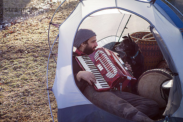Mann spielt Akkordeon für Hund  während er im Zelt auf dem Campingplatz sitzt