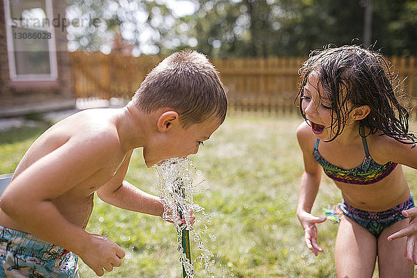 Schwester mit geöffnetem Mund schaut dem Bruder beim Trinken von Wasser durch den Gartenschlauch im Hof zu