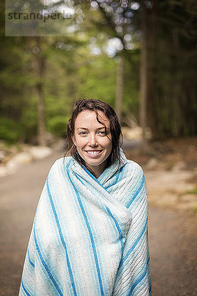 Porträt einer lächelnden  in eine Decke gehüllten Frau  die auf einer Straße im Wald steht
