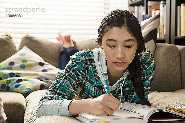 Teenagerin macht Hausaufgaben  während sie zu Hause auf der Couch liegt