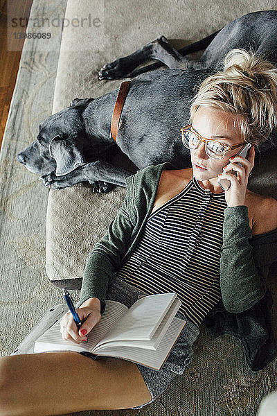 Draufsicht einer Frau  die mit einem Smartphone mit Tagebuch spricht  während sie sich zu Hause auf Dogge entspannt