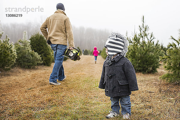 Kleinkind steht auf Grasfeld mit Vater  der eine schnurlose Kettensäge trägt
