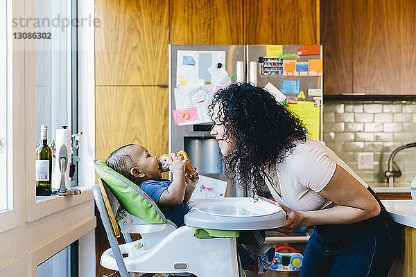 Seitenansicht einer Mutter  die mit ihrem Sohn spielt  der zu Hause in der Küche auf einem Hochstuhl sitzt