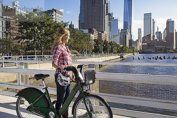 Nachdenkliche Frau mit Fahrrad auf der Brücke mit Blick auf Fluss gegen Stadt