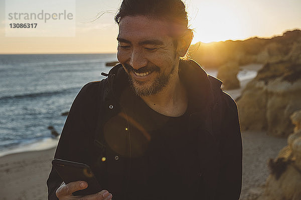 Lächelnder Mann benutzt Smartphone  während er bei Sonnenuntergang am Strand steht