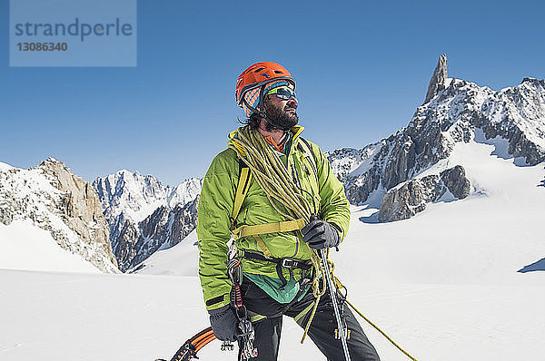 Wanderer mit Kletterausrüstung steht auf schneebedecktem Berg vor strahlend blauem Himmel