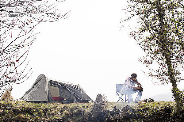 Mann sitzt auf einem Stuhl am Lagerfeuer auf einem Campingplatz auf einem Hügel vor klarem Himmel