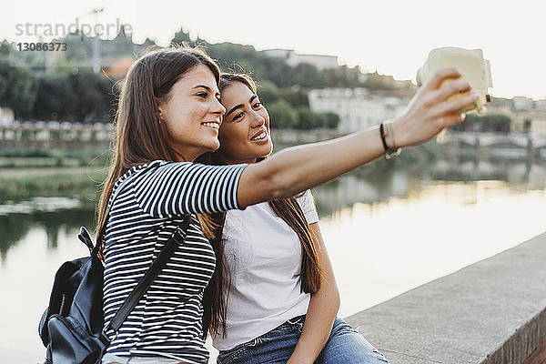 Freunde fotografieren sich selbst mit Sofortbildkamera  während sie auf einer Stützmauer am Fluss Arno in der Stadt sitzen