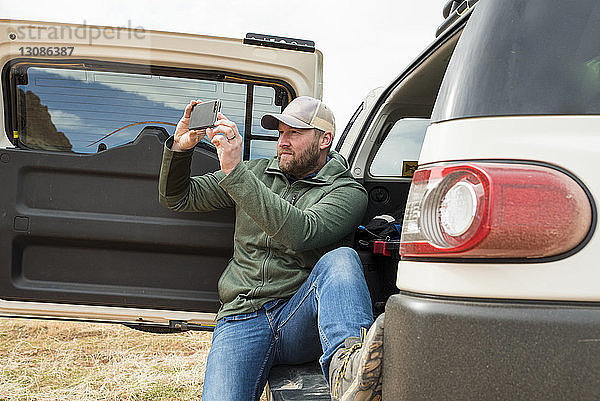 Mann fotografiert mit Smartphone  während er im Kofferraum eines Autos in der Wüste sitzt