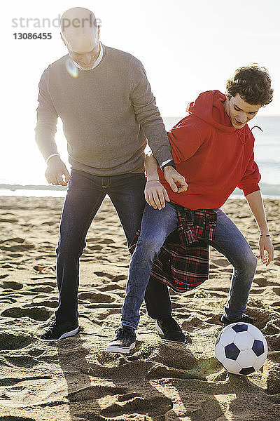 Vater und Sohn in voller Länge beim Fußballspielen am Strand an einem sonnigen Tag