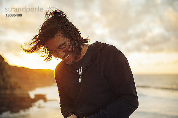 Glücklicher Mann lächelt  während er bei Sonnenuntergang am Strand vor bewölktem Himmel steht