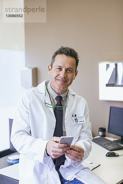 Porträt eines lächelnden Arztes  der ein Smartphone in der Hand hält  während er im Krankenhaus sitzt