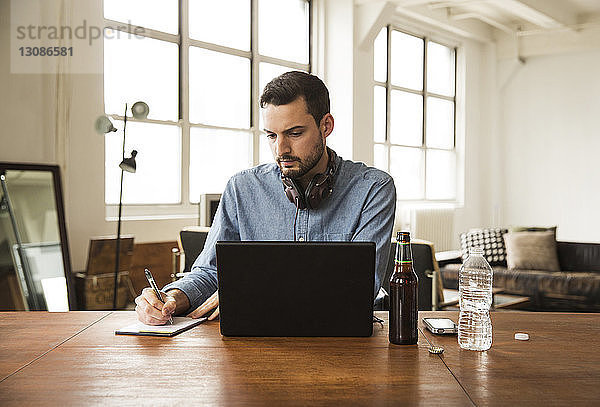 Geschäftsmann schreibt auf einem Notizblock  während er einen Laptop am Tisch im Kreativbüro benutzt