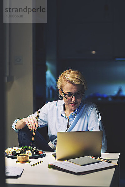 Geschäftsfrau  die nach Essen sucht  während sie am Schreibtisch im Heimbüro einen Laptop benutzt