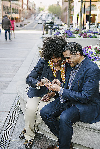 Glückliches Paar benutzt Smartphone  während es in der Stadt auf einem Sitzplatz sitzt