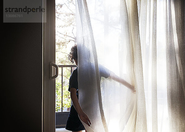 Rückansicht eines Mädchens  das bei Vorhängen am Fenster steht