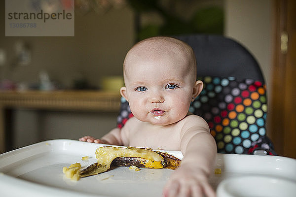 Porträt eines hemdlosen Mädchens mit Banane auf einem Hochstuhl sitzend