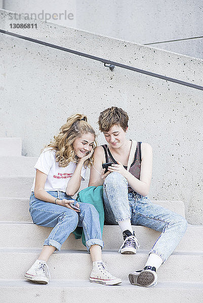 Freundinnen in voller Länge hören Musik  während sie auf Stufen in der Stadt ein Smartphone betrachten