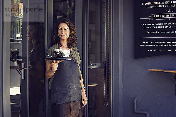 Porträt einer selbstbewussten Besitzerin  die vor dem Café ein Tablett mit Kaffeetasse hält