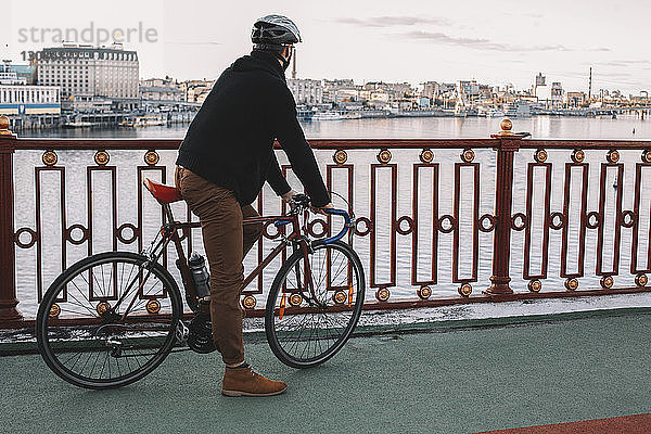 Seitenansicht eines männlichen Radfahrers mit Fahrrad  der auf einer Brücke über den Fluss gegen den Himmel in der Stadt steht