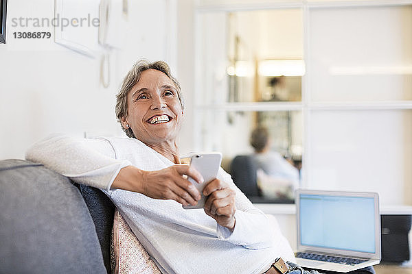 Fröhliche reife Frau hält Smartphone in der Hand  während sie zu Hause auf dem Sofa sitzt