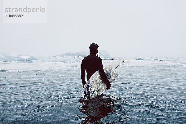 Mann trägt Surfbrett  während er im Winter im eisigen Meer vor klarem Himmel steht