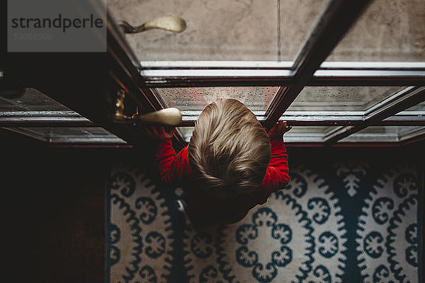 Hochwinkelansicht eines kleinen Jungen  der zu Hause durch eine Glastür schaut