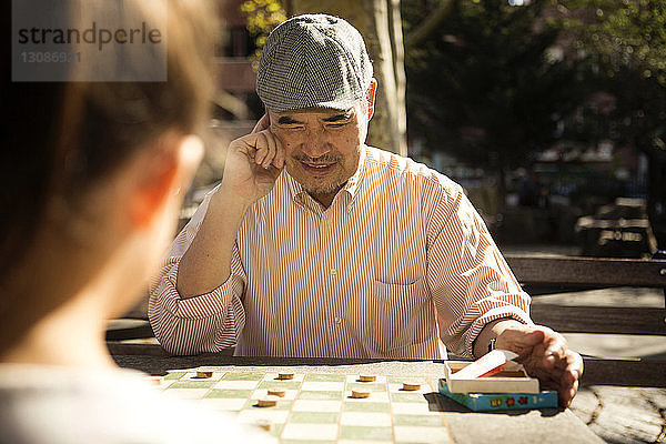 Nachdenklicher älterer Mann spielt Damespiel mit Enkelin am Tisch im Park