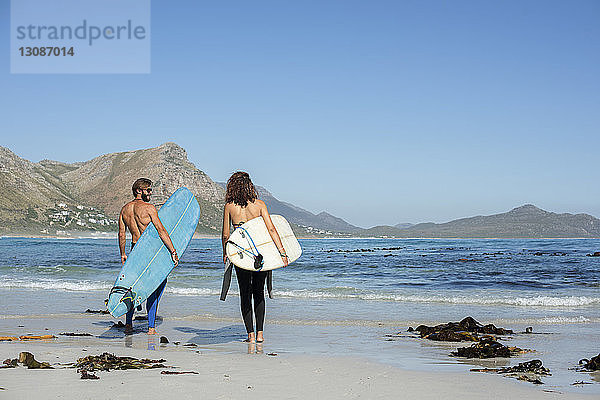 Rückansicht von Freunden mit Surfbrett beim Strandspaziergang am sonnigen Tag
