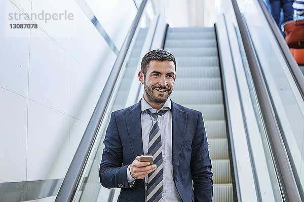 Glücklicher Geschäftsmann hält Smartphone auf Rolltreppe in U-Bahn-Station