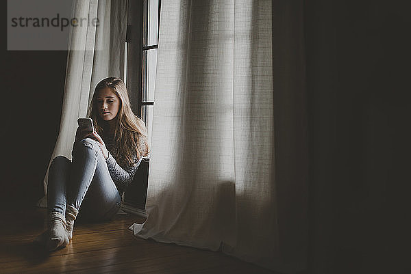 Teenager-Mädchen in voller Länge benutzt ein Smartphone  während sie zu Hause am Fenster sitzt