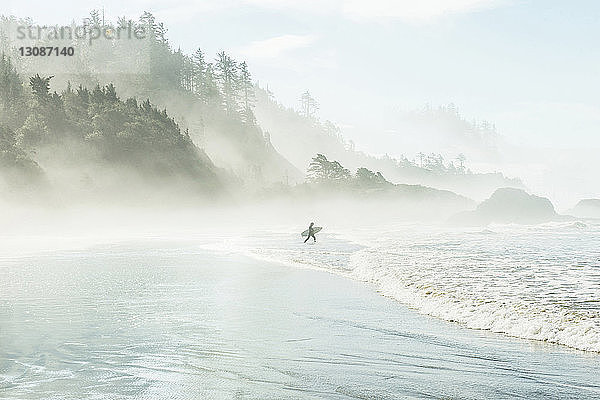 Mann mit Surfbrett  der bei nebligem Wetter am Berg Richtung Meer läuft