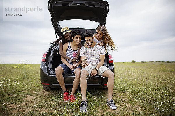 Glückliche Freunde  die ein Smartphone benutzen  während sie im Kofferraum eines Autos auf einem Feld gegen den Himmel sitzen