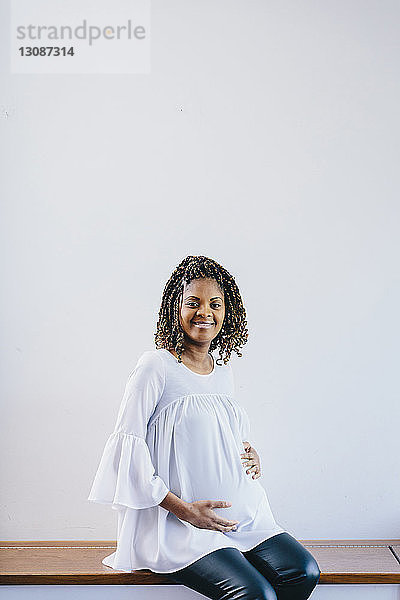 Porträt einer schwangeren Frau  die den Bauch berührt  während sie auf einer Bank vor weißem Hintergrund sitzt