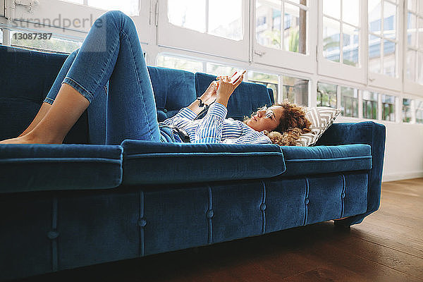 Junge Frau in voller Länge  die ein Smartphone benutzt  während sie zu Hause auf dem Sofa liegt