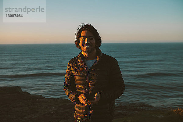 Porträt eines glücklichen Mannes  der ein Smartphone in der Hand hält  während er bei Sonnenuntergang am Meer steht