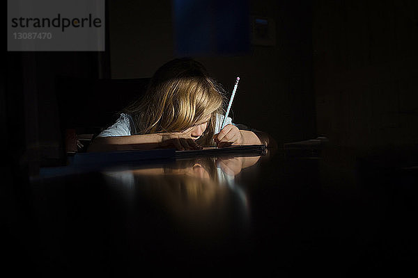 Mädchen schreibt Hausaufgaben  während sie in der Dunkelkammer des Hauses am Tisch sitzt