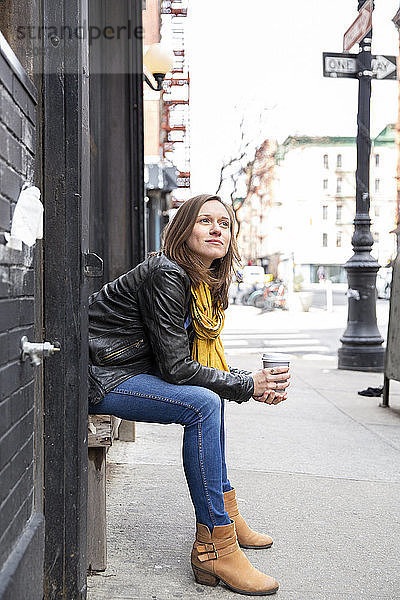 Seitenansicht einer Frau  die Kaffee hält  während sie auf einem Sitz an einem Gebäude in der Stadt sitzt