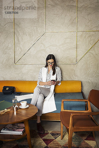 Lächelnde Geschäftsfrau benutzt Smartphone  während sie auf dem Sofa in der Hotellobby sitzt