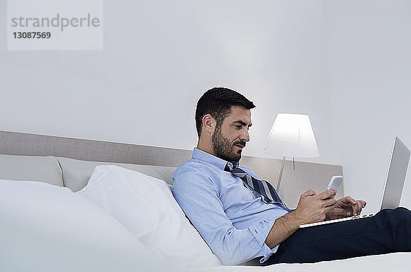 Seitenansicht eines Geschäftsmannes  der ein Smartphone und einen Laptop benutzt  während er im Hotelzimmer auf dem Bett sitzt