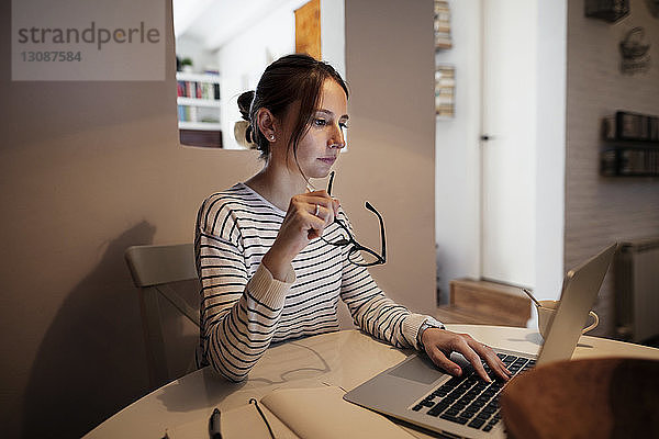 Konzentrierte Frau  die eine Brille hält  während sie am Tisch einen Laptop benutzt