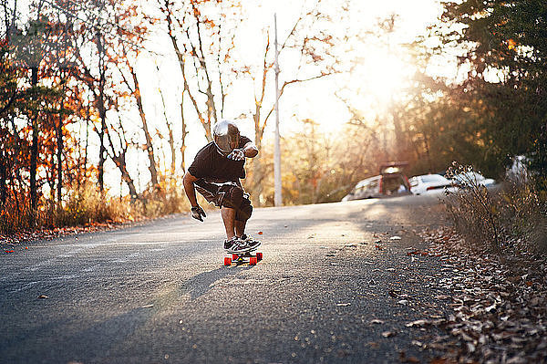 Mann fährt bei Sonnenschein auf der Straße zwischen Bäumen Skateboard