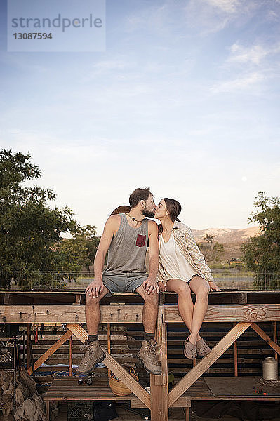 Liebevolles Bauernpärchen küsst sich  während es auf einer Holzkonstruktion auf dem Bauernhof gegen den Himmel sitzt