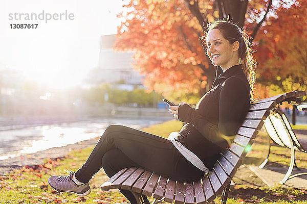 Glückliche Frau hält Handy in der Hand  während sie auf einer Parkbank sitzt